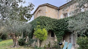 Un bâtiment en pierre avec un toit de tuiles, des murs couverts de lierre et une petite fenêtre. On aperçoit deux fauteuils de jardin verts au premier plan.
