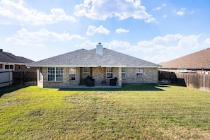 A single-story house with a sloped roof, a front porch with two chairs, and a fenced backyard.