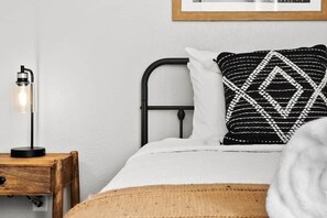 A bed with a white mattress, a black metal headboard, a patterned pillow, a bedside table with a lamp, and a wooden nightstand.