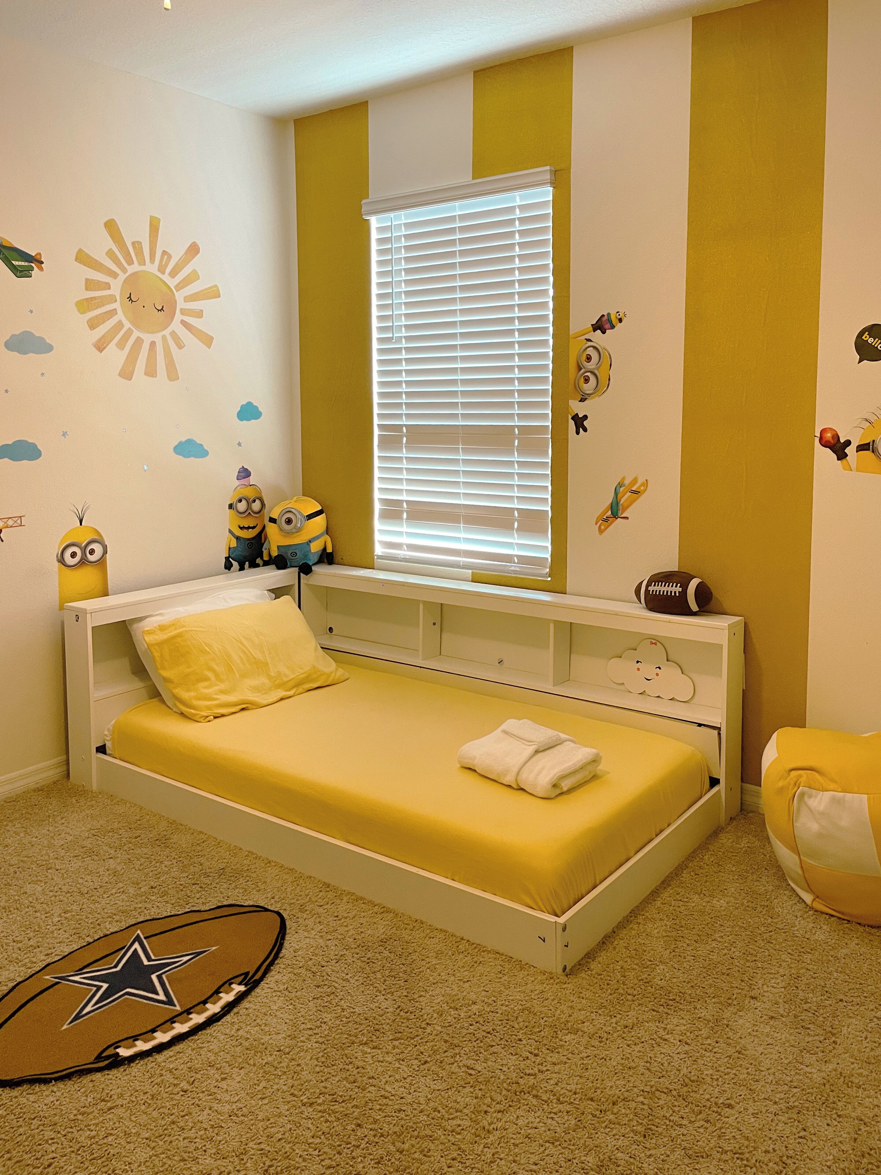 A child's bedroom with a bed, a window with blinds, and wall decorations including a sun, clouds, and minions.