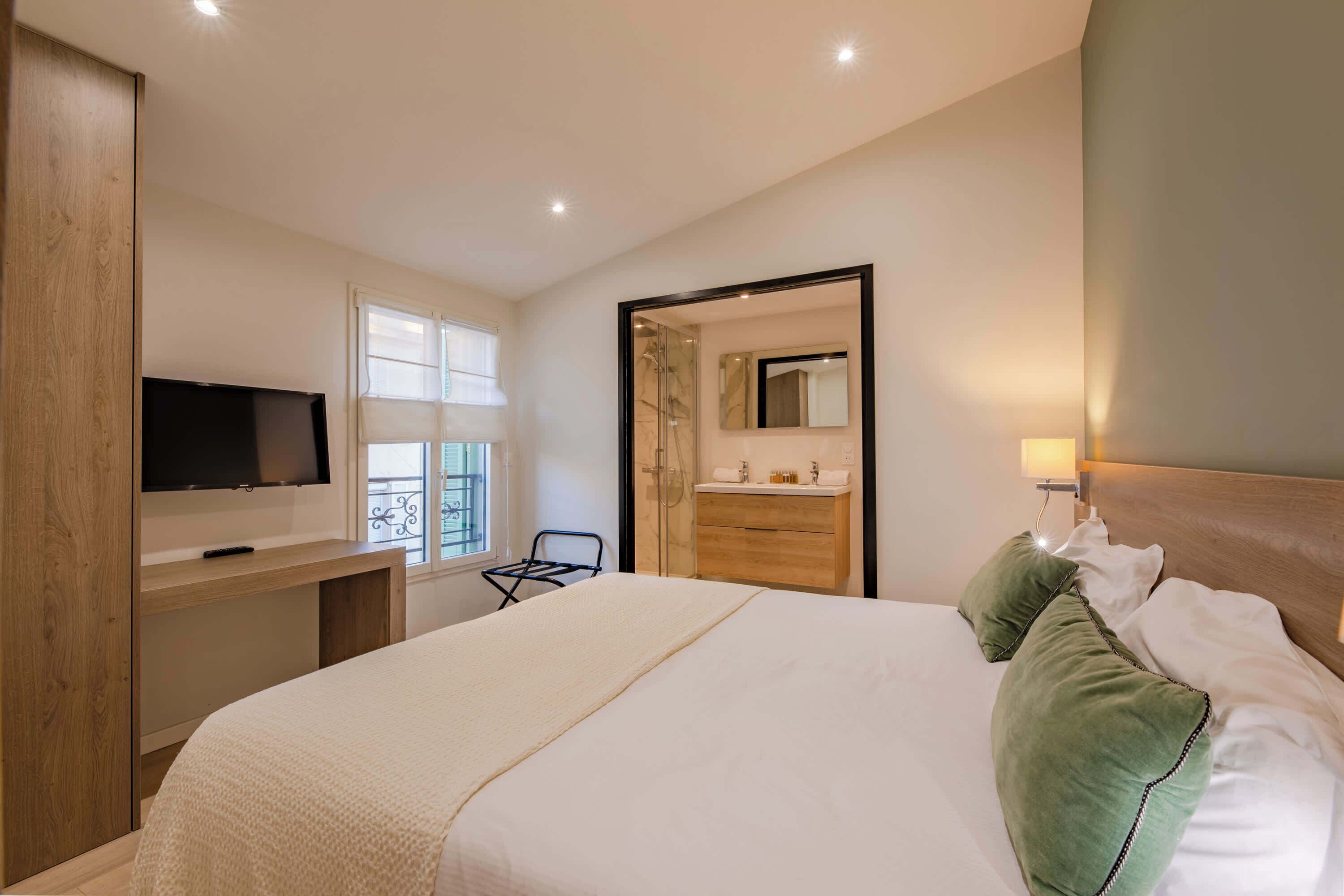 Une chambre d’hôtel avec un lit, un téléviseur posé sur un bureau en bois, une salle de bain avec un miroir et une fenêtre avec des stores.