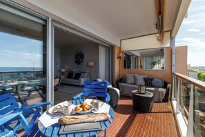 A balcony with a table set for breakfast, blue chairs, a sofa, and a view of the sea.