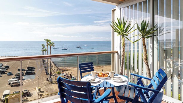 Un balcón con una mesa preparada para dos personas, con vista a una playa con palmeras y barcos.