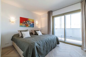 A bedroom with a large bed, two wall-mounted lights, a painting, and a view of the outside through a large window.