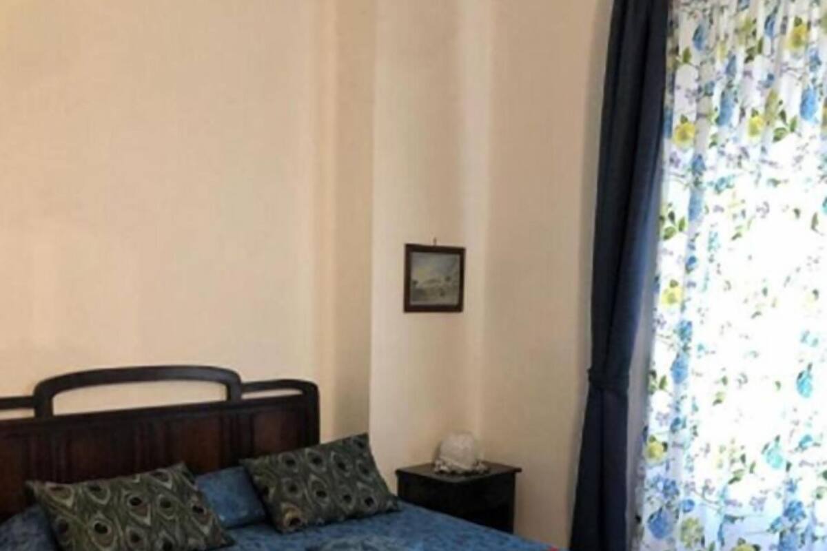 A neatly made bed with a blue bedspread and decorative pillows, a small bedside table with a lamp, and a window with floral curtains.