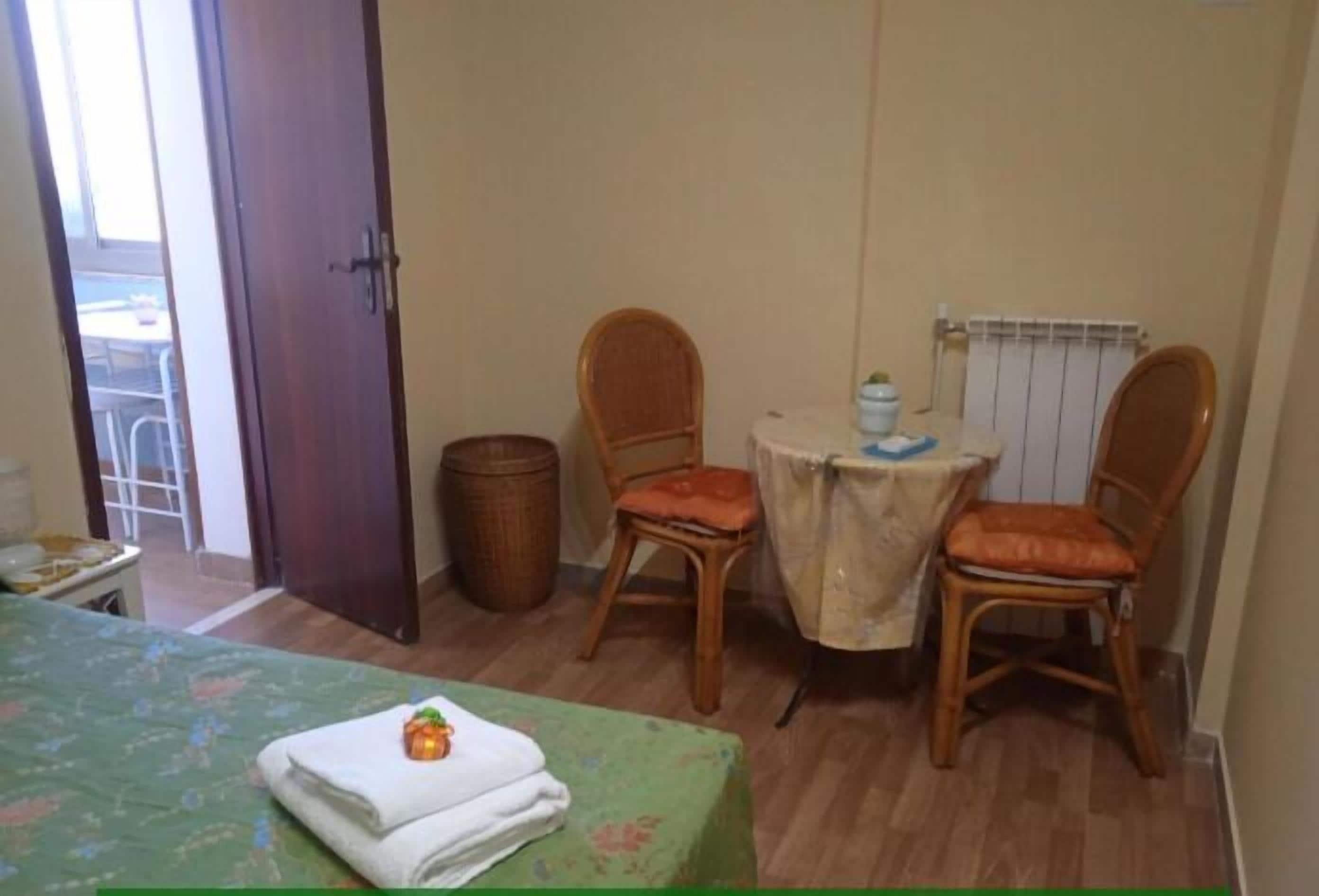 Une petite pièce comprenant un lit, deux fauteuils en osier, une petite table recouverte d’une nappe, un radiateur et une porte donnant sur un espace extérieur.