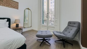 Un dormitorio con cama, una silla, un reposapiés, un espejo y una ventana con vistas a edificios.
