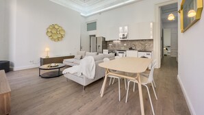 Una sala moderna con un sofá, una mesa redonda de centro y una mesa de comedor con sillas. Se ve una cocina al fondo con armarios blancos y una salpicadura de mármol.