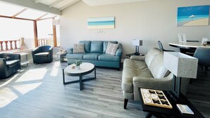 Amplia sala de estar con un sofá azul, un sofá color beige y una mesa de centro redonda. Se observa una mesa de comedor con sillas y una lámpara. La habitación cuenta con un ventanal con vistas al exterior.