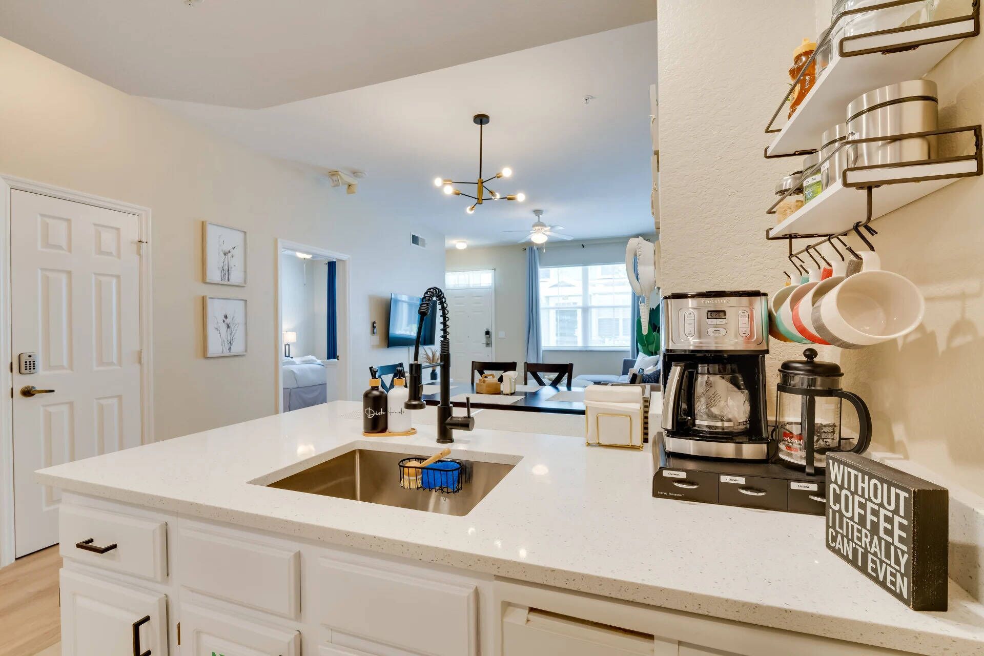 A modern kitchen with a white countertop, stainless steel sink, and a coffee maker. There is a wall-mounted shelf with dishes and a sign that reads "WITHOUT COFFEE I CANNOT EVEN".