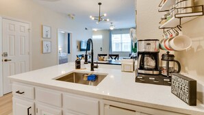 A modern kitchen with a white countertop, stainless steel sink, and a coffee maker. There is a wall-mounted shelf with dishes and a sign that reads "WITHOUT COFFEE I CANNOT EVEN". - Family Escape Near Disney | Resort Amenities (Kissimmee)