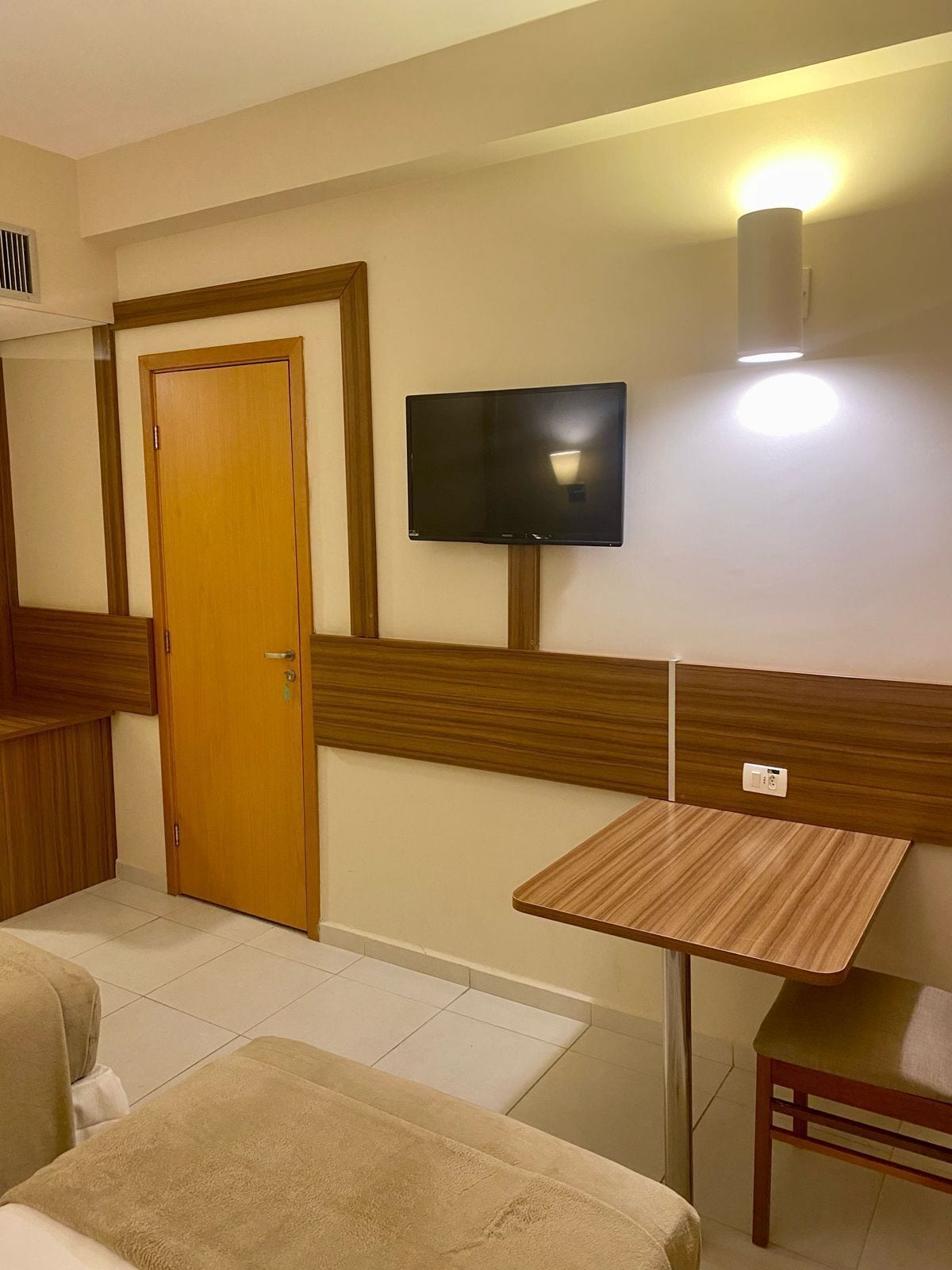 A hotel room with a wooden door, a flat-screen TV mounted on the wall, a small wooden table, and a bed with a beige blanket.