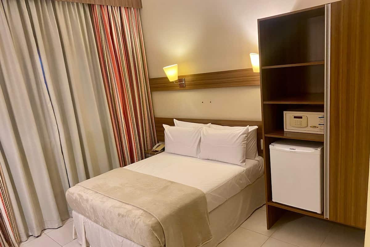 A hotel room with a bed, a bedside table with a lamp, a small refrigerator, and a striped curtain.
