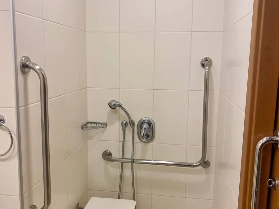 Une petite cabine de douche aux murs recouverts de carreaux blancs, équipée d’un pommeau de douche à main, de barres d’appui et d’un flexible de douche à main.