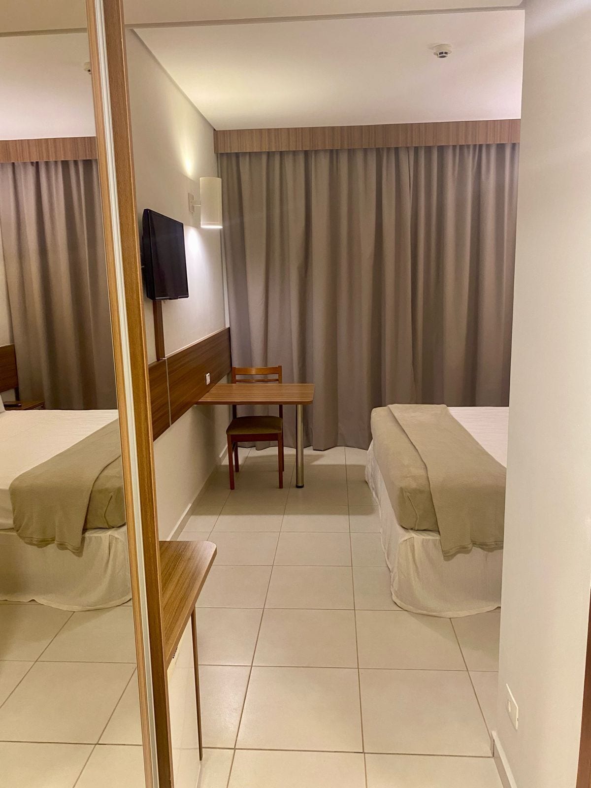 A hotel room with two single beds, a wooden desk, a chair, a television mounted on the wall, and a window with curtains.