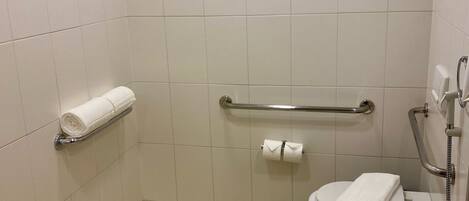 A small, clean bathroom with white tiles, a toilet, a handrail, and a shower with a handheld showerhead.