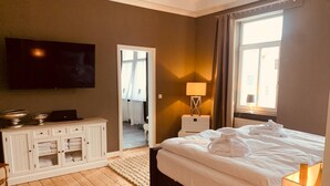 Habitación de hotel con una cama grande, televisor montado en la pared, mesita de noche con lámpara y vistas al exterior a través de una ventana.