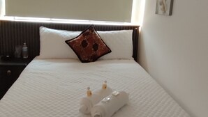 Een netjes opgemaakt bed met witte lakens en een decoratief kussen, een nachtkastje met een fles erop, en wanddecoratie.