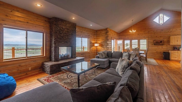 A spacious living room with a stone fireplace, large windows, and a high ceiling. - Goat Mountain Cabin: Mountain Views & Big Sky Comfort in Livingston (Livingston)