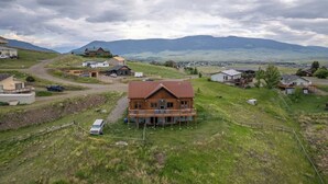 A large wooden house with a porch, surrounded by a rural landscape with hills and a small town in the background. - Goat Mountain Cabin: Mountain Views & Big Sky Comfort in Livingston (Livingston)
