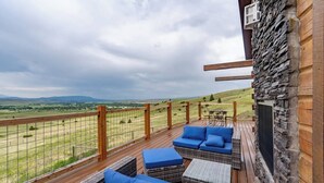 A wooden deck with wicker furniture and a stone wall, overlooking a grassy field and distant hills. - Goat Mountain Cabin: Mountain Views & Big Sky Comfort in Livingston (Livingston)