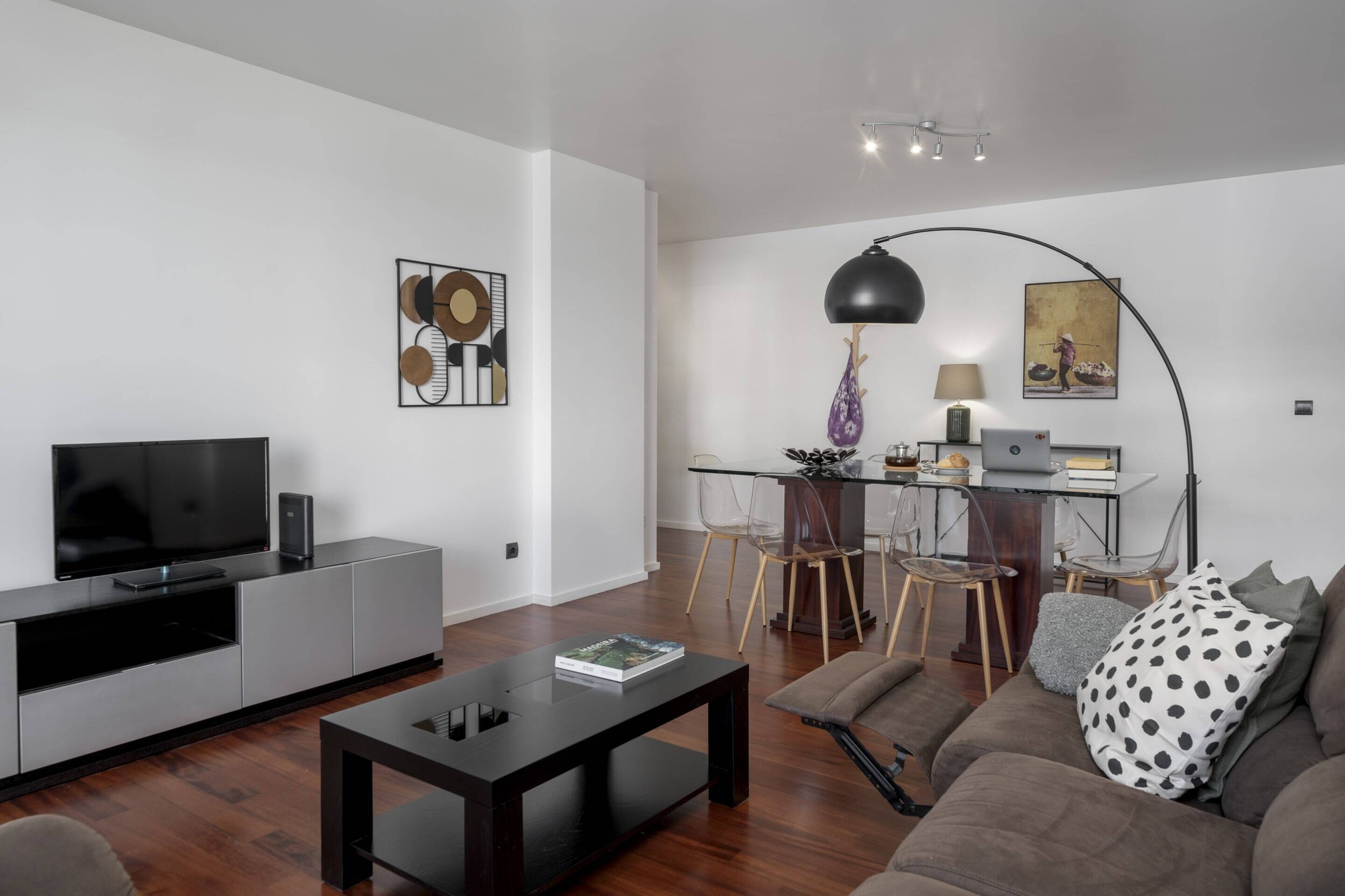 A modern living room with a flat-screen TV, a coffee table, and a sofa. There is a dining area with a table and chairs, and a lamp.