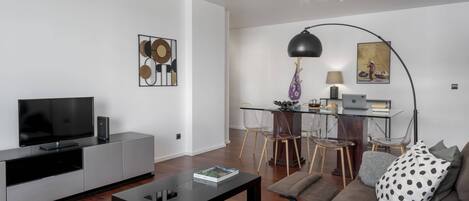 A modern living room with a flat-screen TV, a coffee table, and a sofa. There is a dining area with a table and chairs, and a lamp.