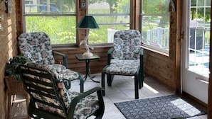 L’intérieur d’un chalet en bois est doté d’un ventilateur de plafond, de deux fauteuils à bascule, d’une table et de grandes fenêtres qui offrent une vue sur la verdure environnante.