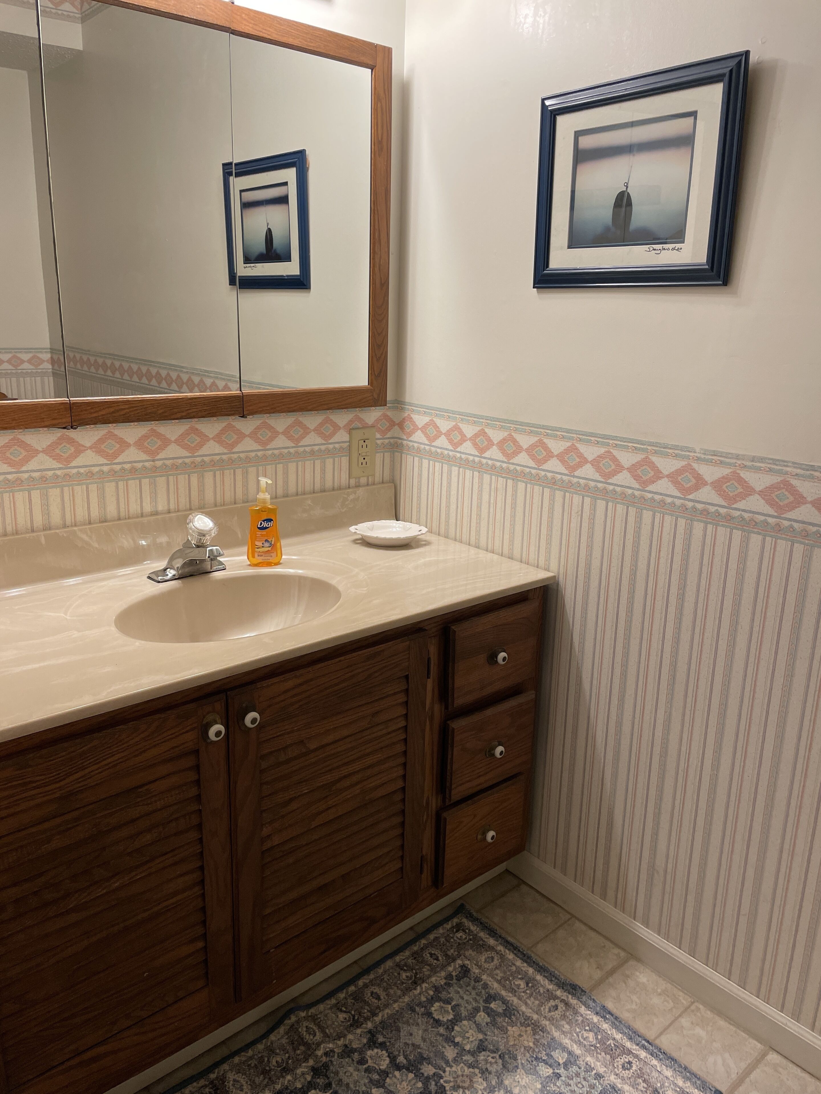 Une salle de bain avec un meuble-lavabo en bois, un évier, un miroir, un tableau encadré au mur et un tapis à motifs.