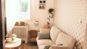 Ein gemütliches Wohnzimmer mit einer Couch, einem Korbstuhl, einem kleinen Tisch mit Pflanze und einem Fenster mit Vorhängen.