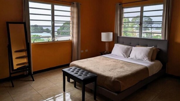 Habitación de hotel con cama, mesita de noche, lámpara, ventilador de techo y ventanas amplias con cortinas.