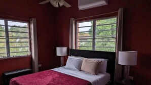 Habitación de hotel con una cama grande, dos lámparas de noche, un banco y ventanas con vistas a zonas verdes.