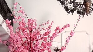 A round table with a vase of pink flowers, a black upholstered sofa, and a chandelier in the background.