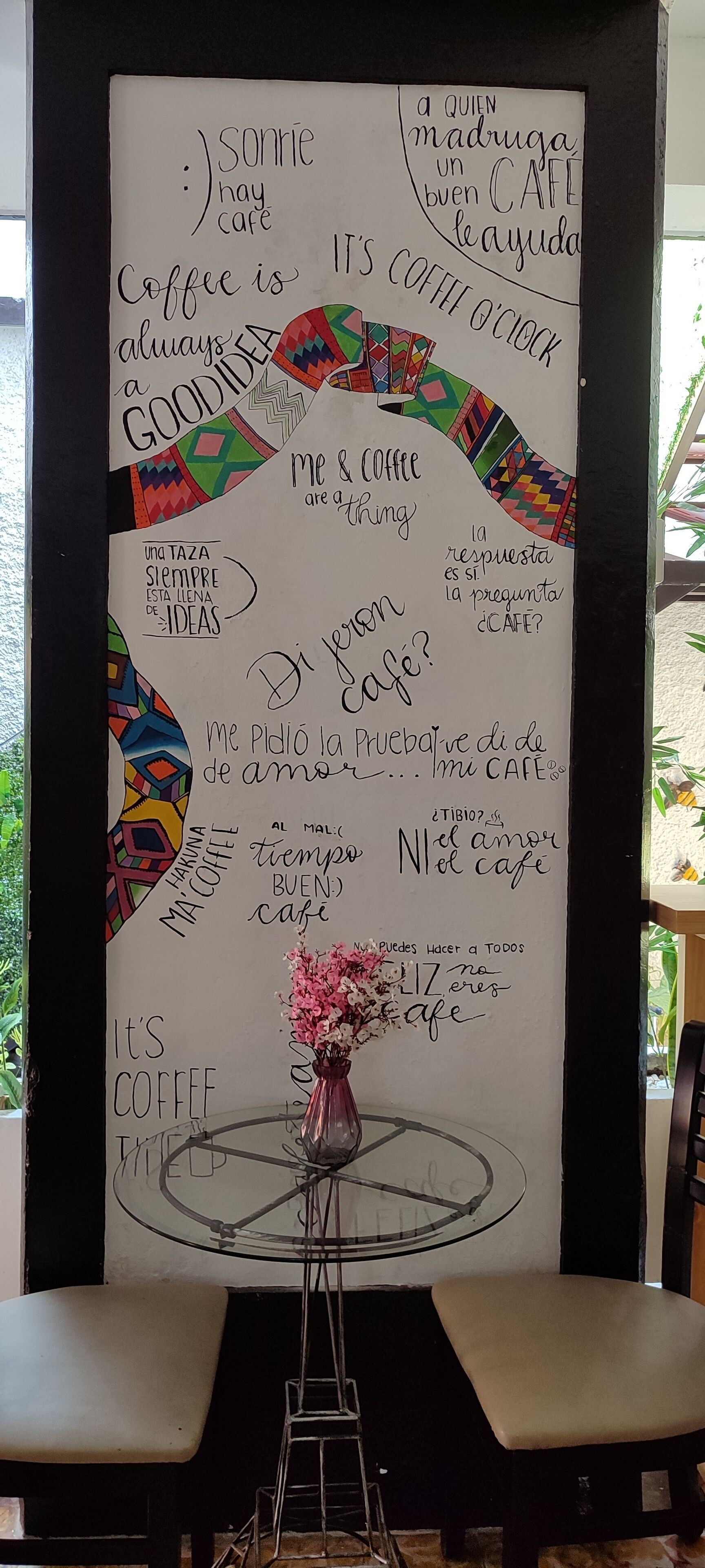 A wall with various handwritten phrases and doodles, including 'Coffee is always a good idea' and 'It's coffee o'clock.' A glass table with a vase of flowers is in front of the wall.