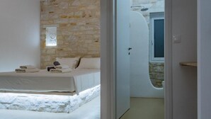 A bedroom with a stone wall, a bed with white linens, and a bathroom with a shower. - Aegean Feelings - Luxury Villa Calm with Private Pool close to Piso Livadi Beach (Piso Livadi)
