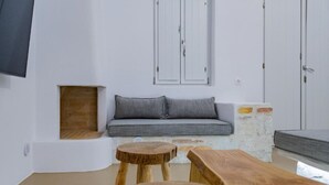 A minimalist living room with a grey sofa, two wooden stools, and a white wall with a small window and a TV mounted above. - Aegean Feelings - Luxury Villa Calm with Private Pool close to Piso Livadi Beach (Piso Livadi)