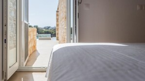 A bedroom with a bed, an open door leading to a balcony, and a stone wall. - Aegean Feelings - Luxury Villa Calm with Private Pool close to Piso Livadi Beach (Piso Livadi)