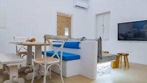 A modern living room with a white sofa, a dining table with chairs, and a television mounted on the wall. - Aegean Feelings - Luxury Villa Calm with Private Pool close to Piso Livadi Beach (Piso Livadi)