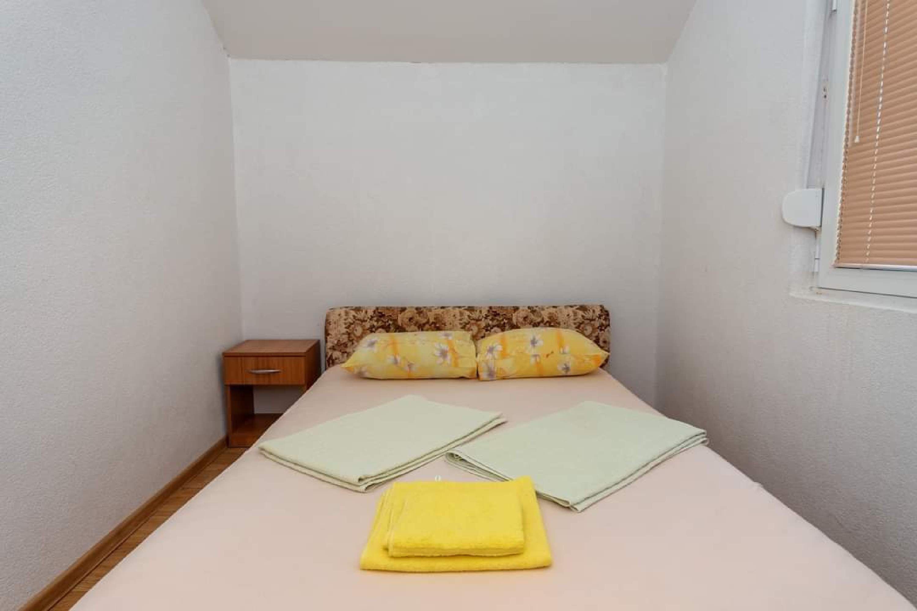 A small, neatly made bed with a floral headboard, a wooden nightstand, and a window with blinds.