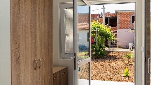 Una habitación con puerta corredera de vidrio que da a un jardín con un árbol pequeño y un edificio de ladrillo al fondo.