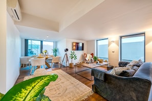 A spacious living area with a sofa, coffee table, and dining table set up. There are potted plants and a large window providing natural light.
