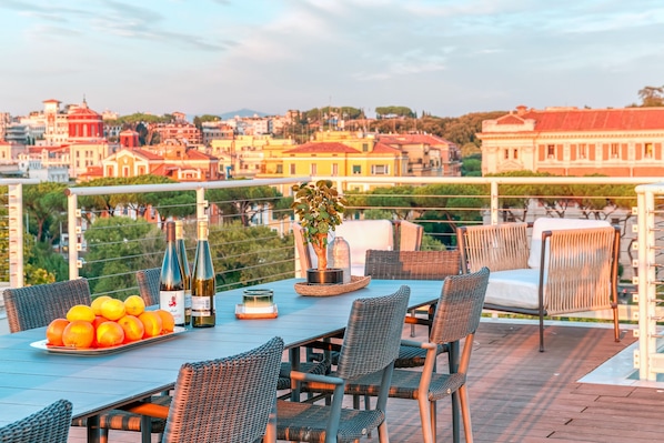 A terrace with a table set for dining, featuring wicker chairs, a bottle of wine, and a potted plant.