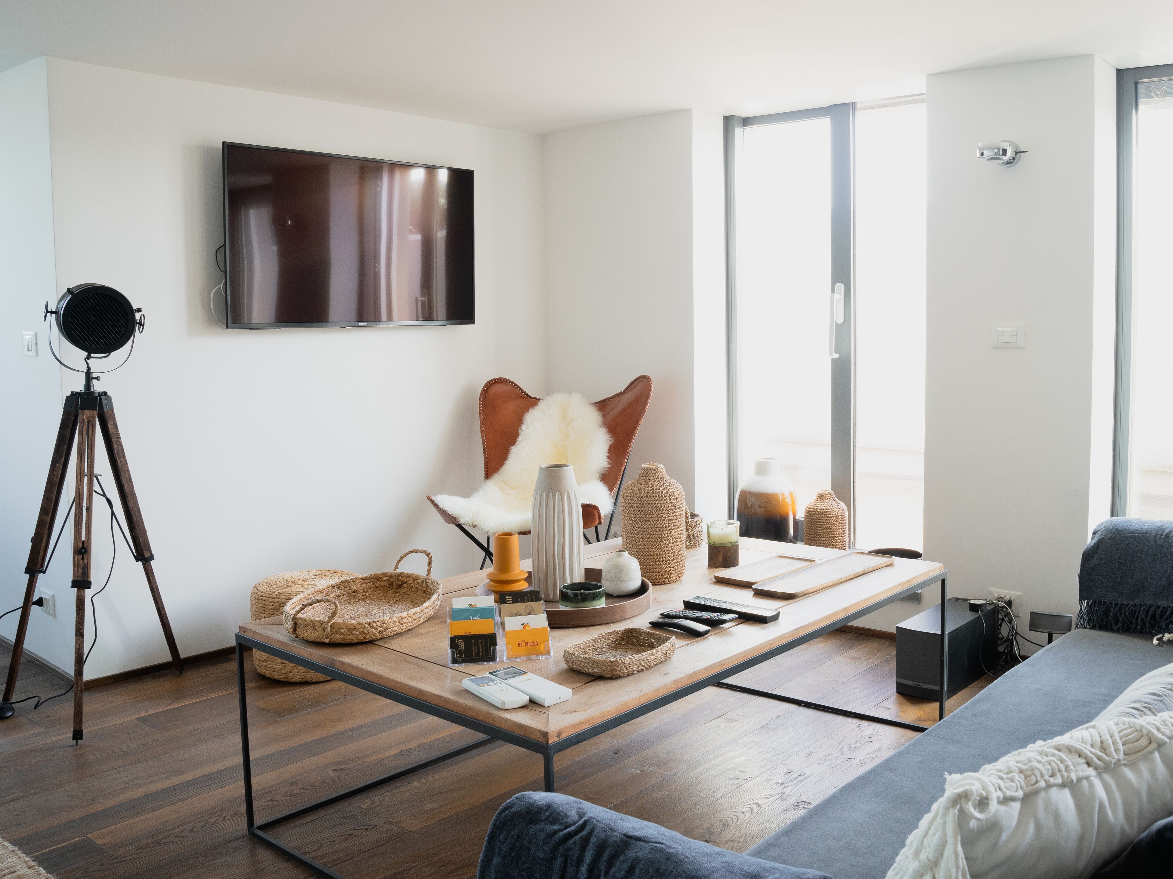 Una sala de estar moderna con un televisor de pantalla plana grande montado en la pared, un trípode, un sillón de cuero, un sofá y una mesa de centro con varios objetos encima.
