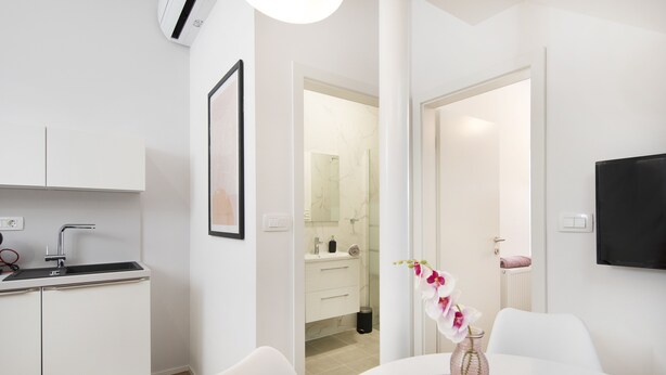 Een moderne keuken met witte kasten, een zwart aanrecht en een spoelbak. Er is een eethoek met een witte tafel en stoelen, en een vaas met bloemen.