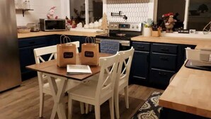 Eine moderne Küche mit dunkelblauen Schränken, einer Holz Arbeitsplatte und einer zentralen Kochinsel mit weißen Stühlen. Auf der Insel stehen zwei Papiertüten und ein kleiner Tisch mit einem Laptop.