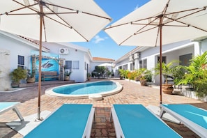 A swimming pool area with lounge chairs and umbrellas, surrounded by potted plants and a building with a mural. - Family-Friendly 1BR w/ Shared Pool, Noord (Noord)