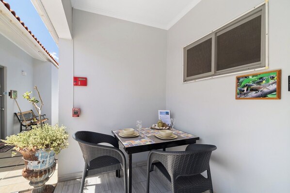 A small dining area with a table set for two, two wicker chairs, a potted plant, and a framed picture on the wall.