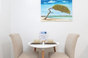 A dining table with two chairs, a menu stand, and a painting of a tree on a beach. - Family-Friendly 1BR Retreat w/ Pool & Beach Access (Noord)