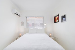 Ein Schlafzimmer mit einem Bett, weißen Wänden, einem Fenster mit Jalousien, einer Klimaanlage, zwei Nachttischlampen und zwei gerahmten Bildern.