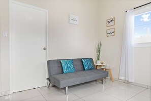 Un salon moderne avec un canapé gris, deux coussins décoratifs, une petite table d’appoint et une fenêtre avec des rideaux.